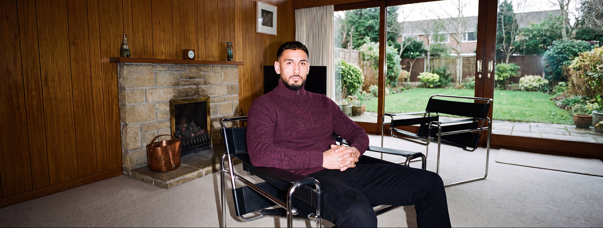 A man wearing a burgundy turtleneck sweater and black trousers sits in a modern chair inside a mid-century modern living room with wood-paneled walls, a stone fireplace, and large windows overlooking a green garden.