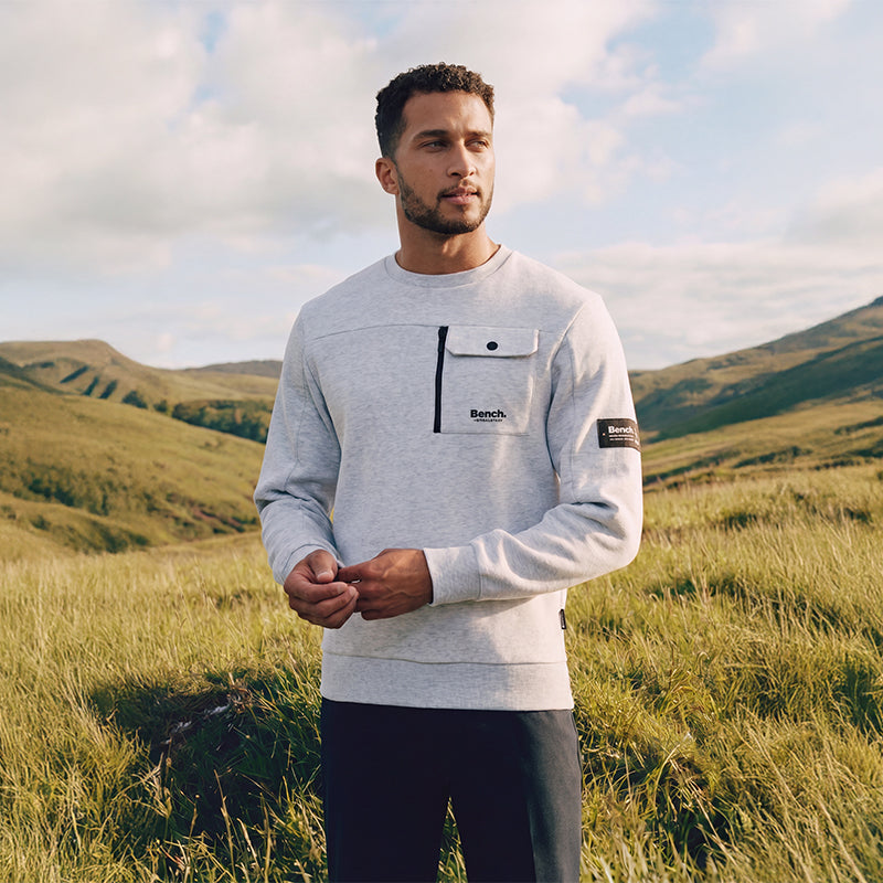 Man wearing a gray sweater with Bench logo in a grassy field with mountains in the background