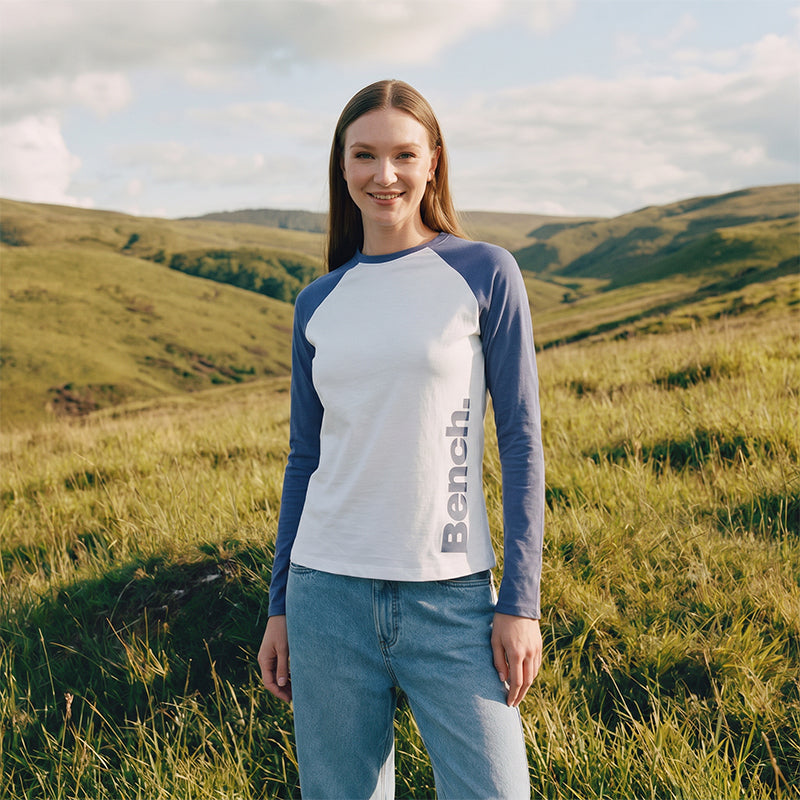 Woman wearing a Bench long-sleeve shirt in a grassy field with hills in the background